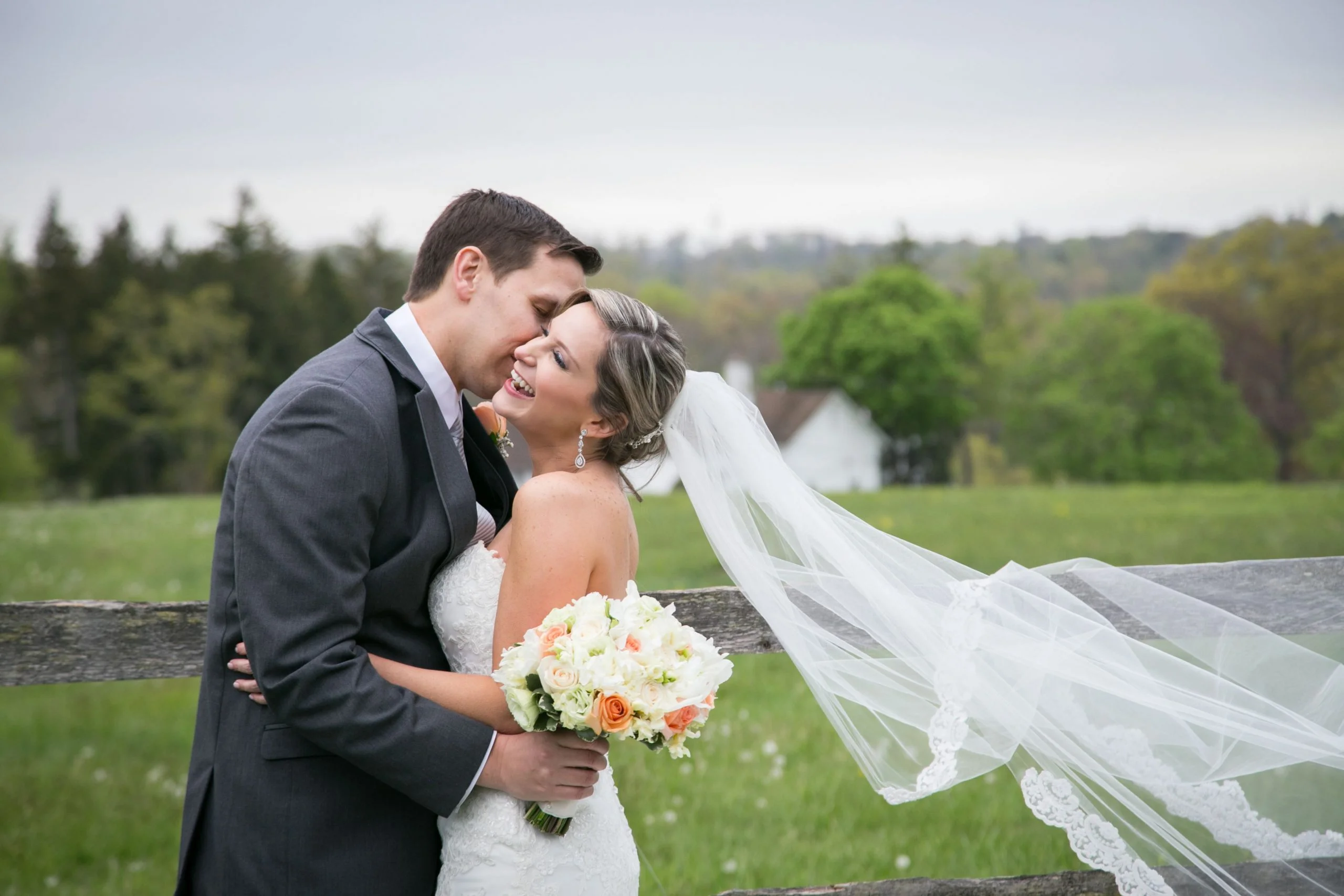 A bride and groom kiss in front of a fence in a field.