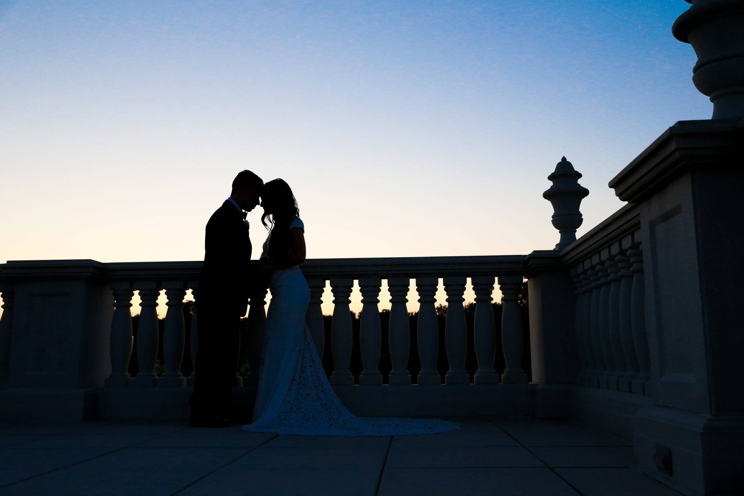 A bride and groom are silhouetted on a balcony at sunset.