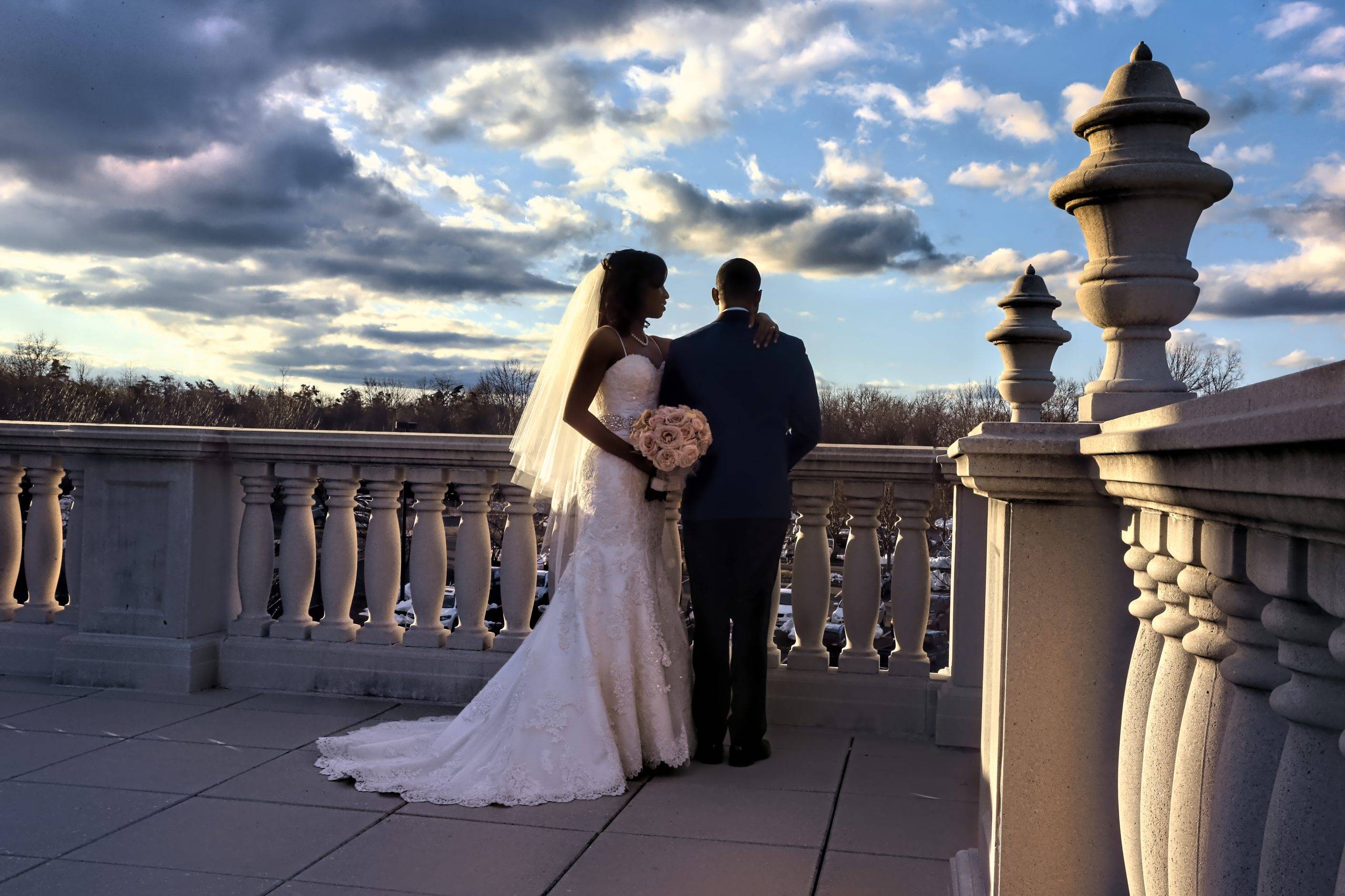 A bride and groom standing on a balcony overlooking the sky.