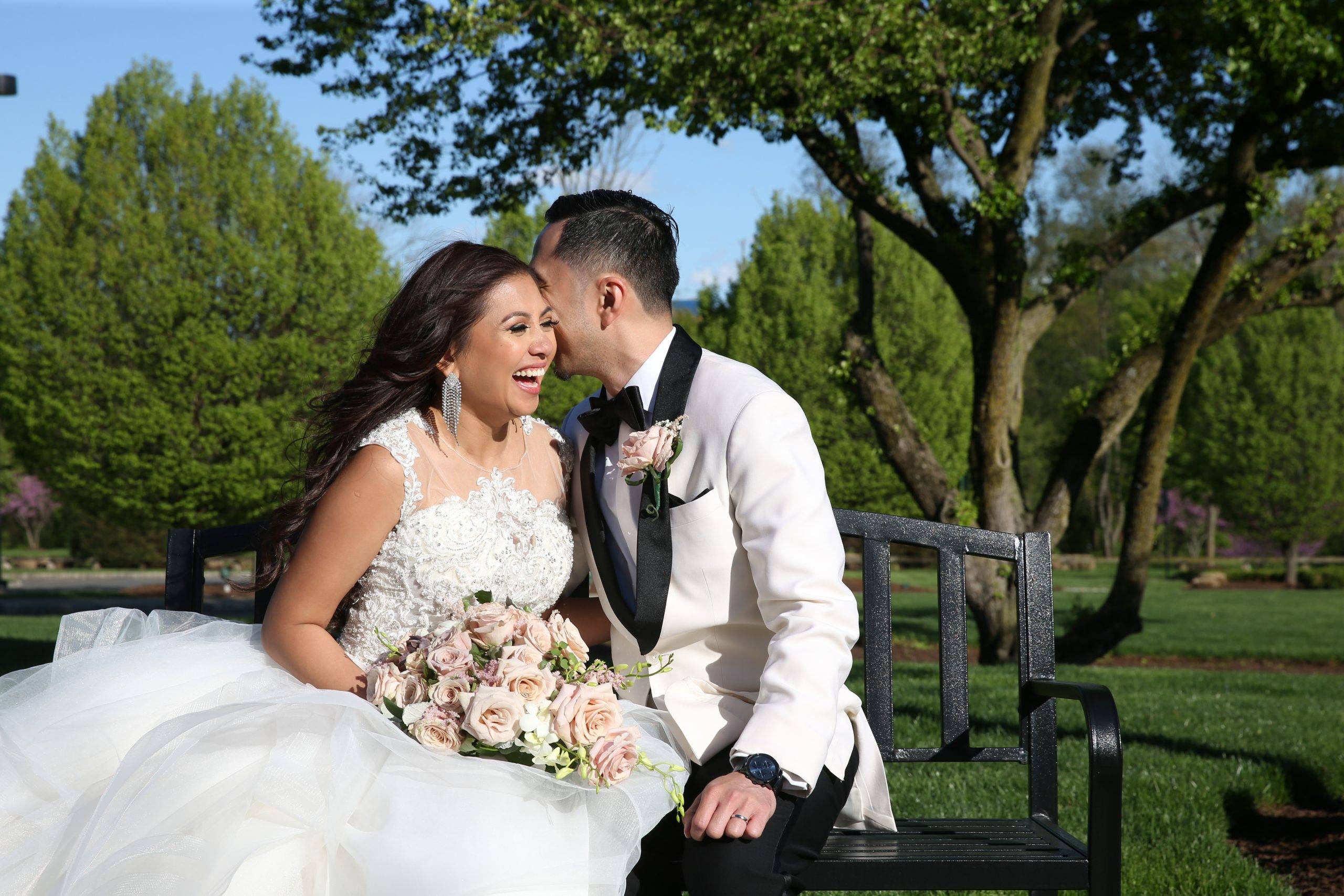A bride and groom kissing on a bench in a park.