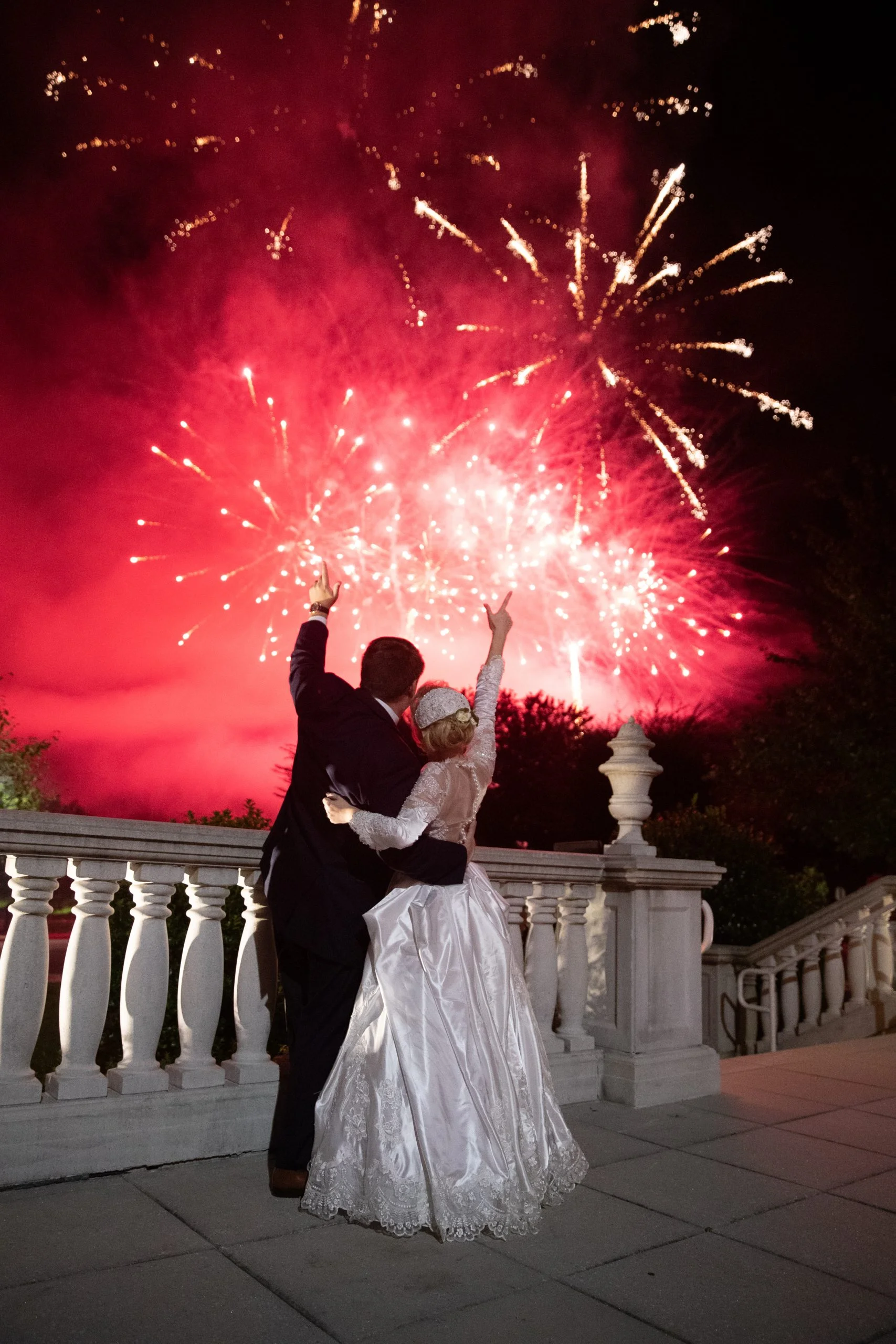 A bride and groom standing on a balcony with fireworks in the background.
