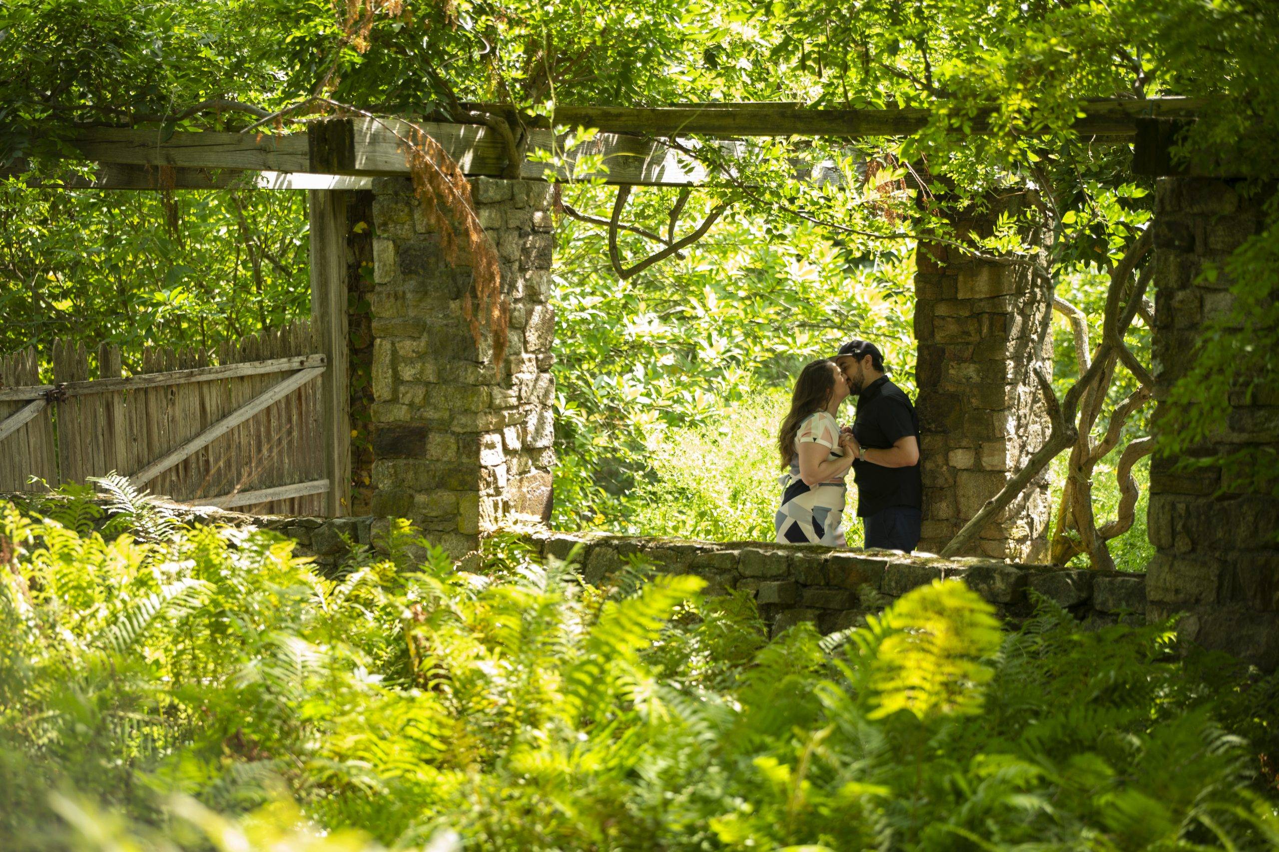 A couple kisses under a stone archway in the woods.
