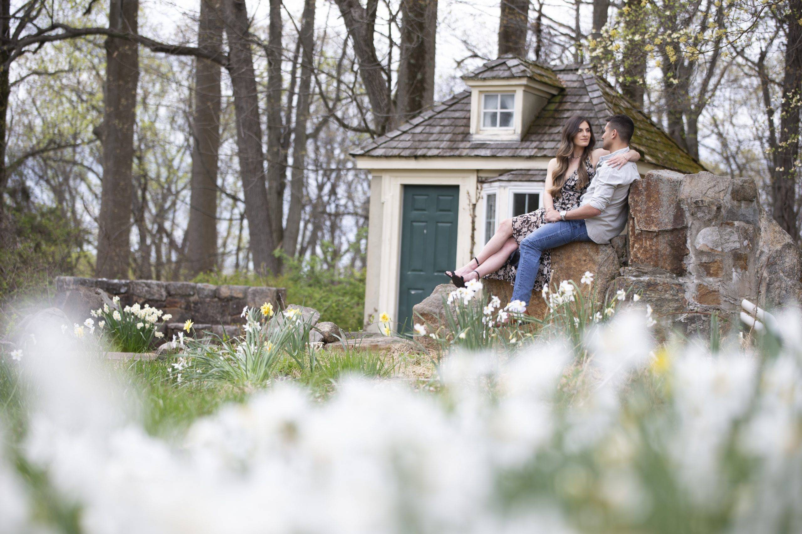 A couple sits on top of a rock in front of a house with daffodils.