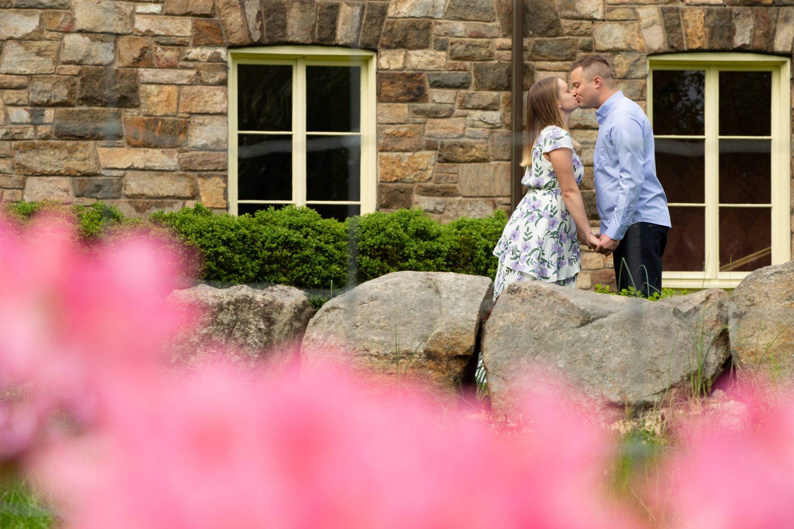 A couple kisses in front of a stone building with pink flowers.