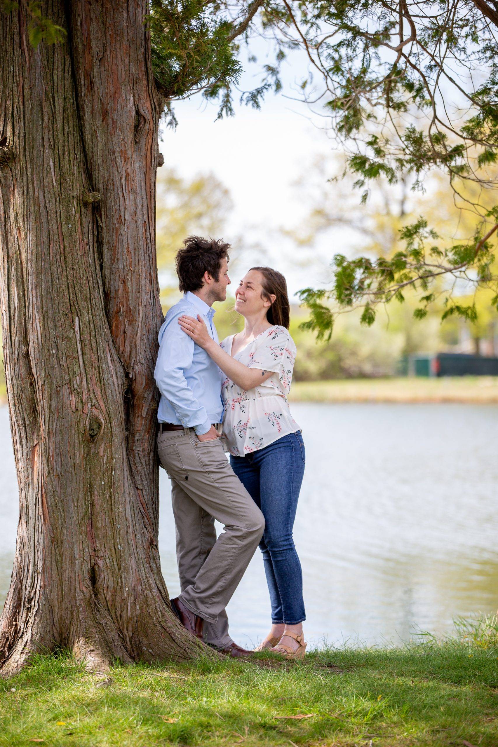 An engaged couple leaning against a tree near a lake.
