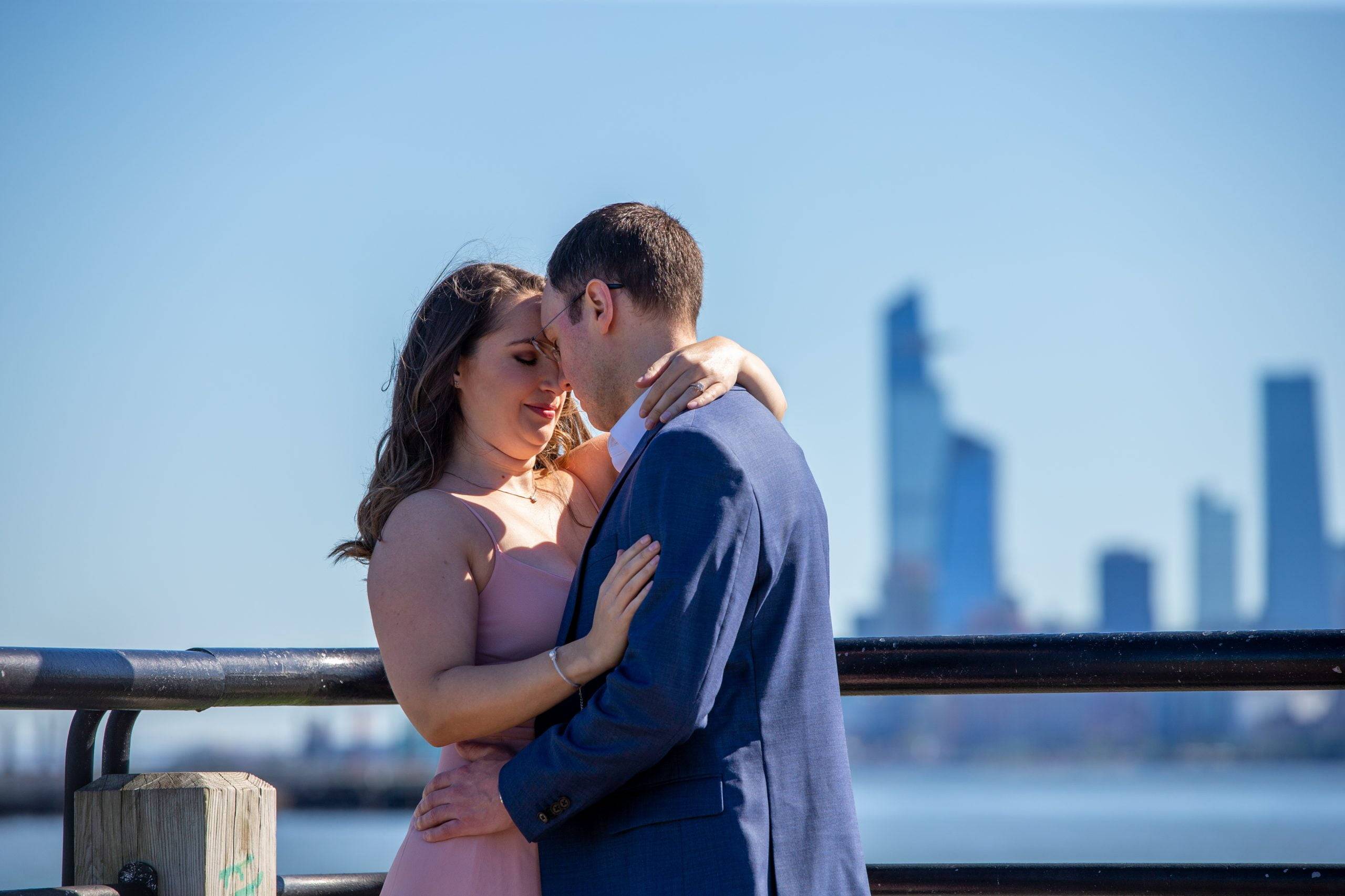 A couple embraces on a pier with the city skyline in the background.
