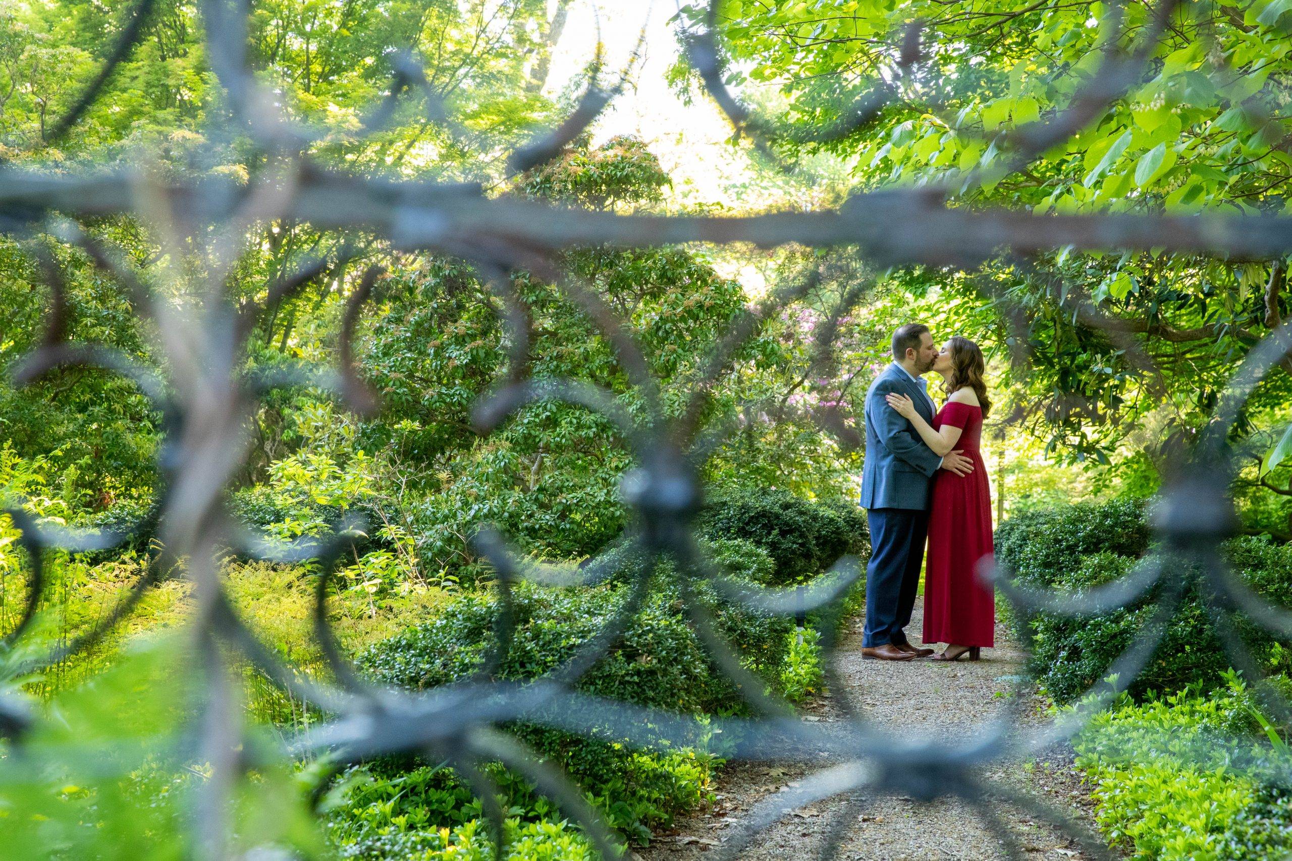 A couple kisses in front of a fence in a garden.
