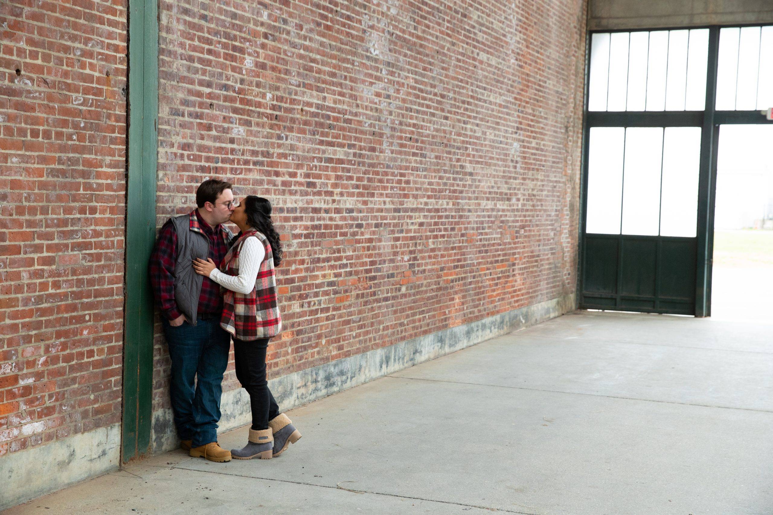 A couple kisses in front of a brick wall.
