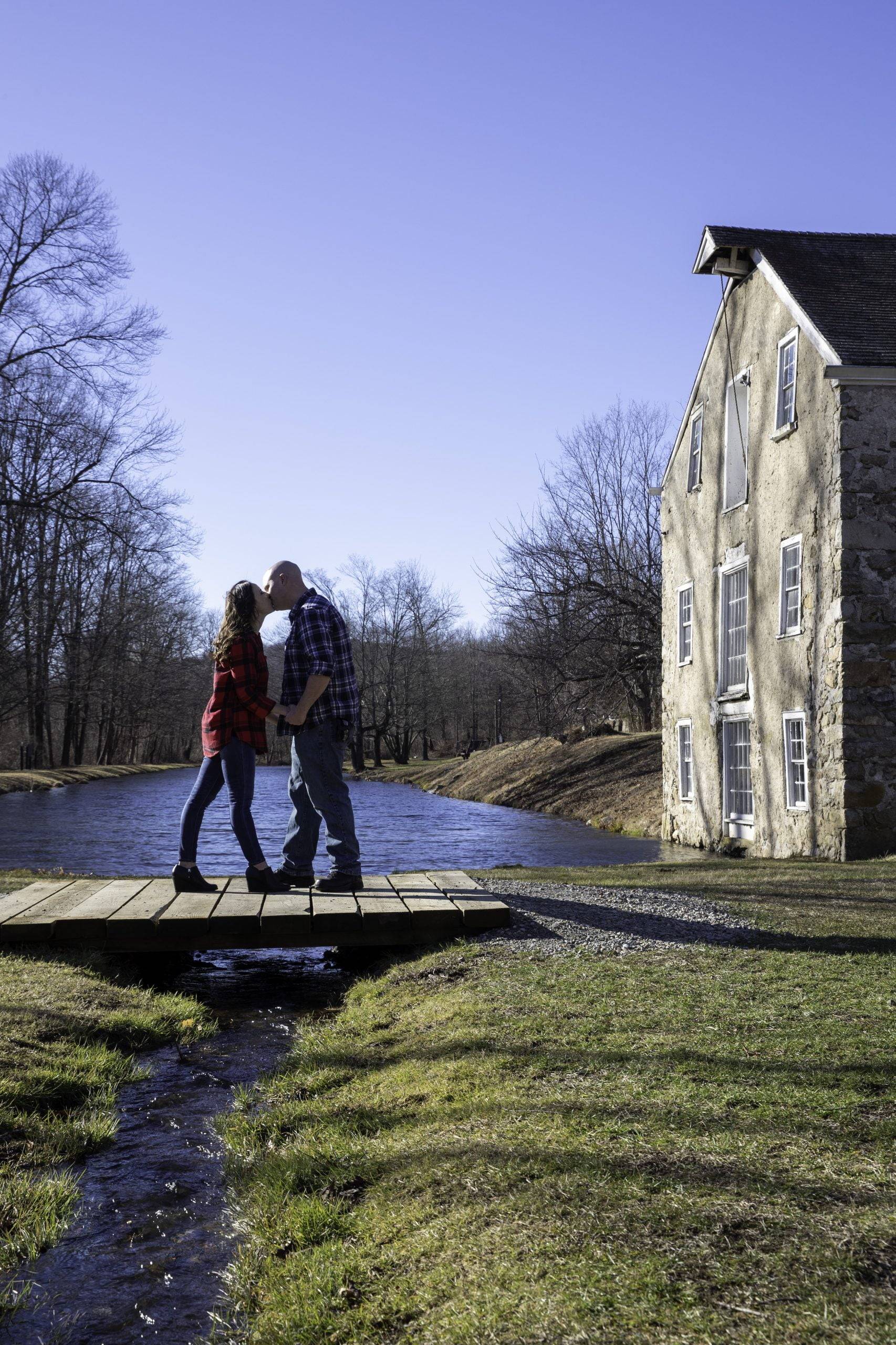 A couple standing on a bridge in front of an old mill.