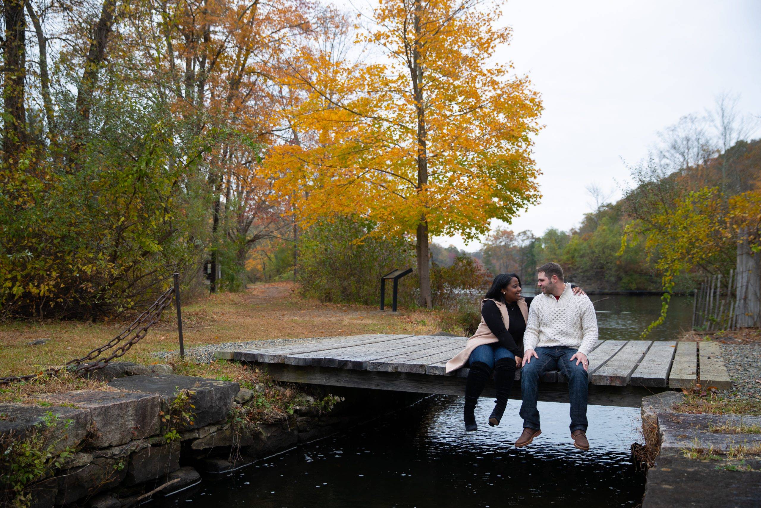 A couple sits on a wooden bridge in the fall.