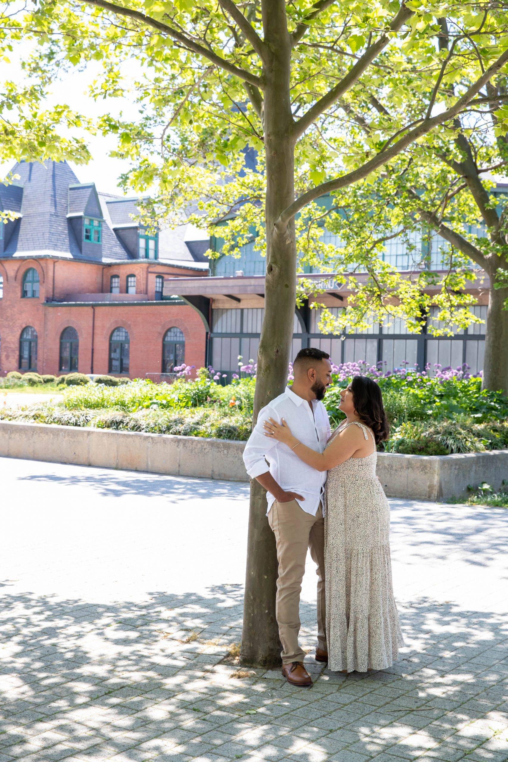 A couple embraces under a tree in front of a brick building.
