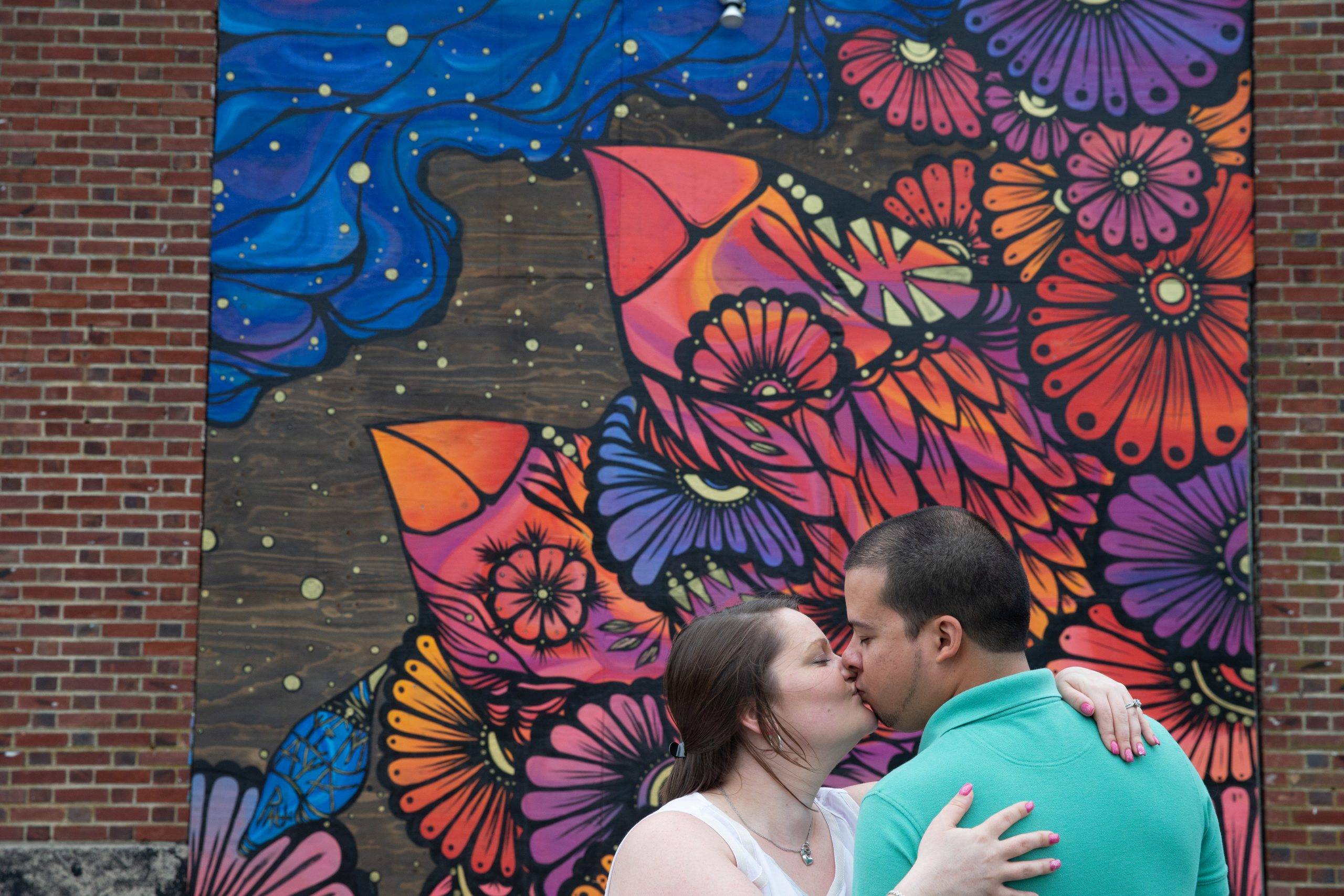 A couple kisses in front of a colorful mural.