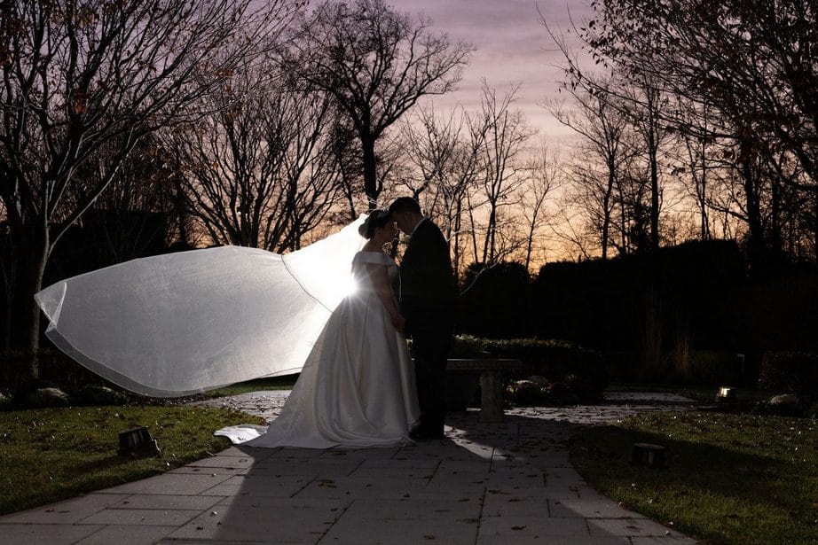 A bride and groom standing holding hands on a picturesque courtyard garden path with trees at sunset