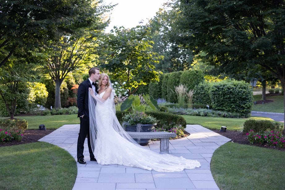 Bride and groom pose for a photo on a garden path as he kisses her head and she has her veil draped over her hand