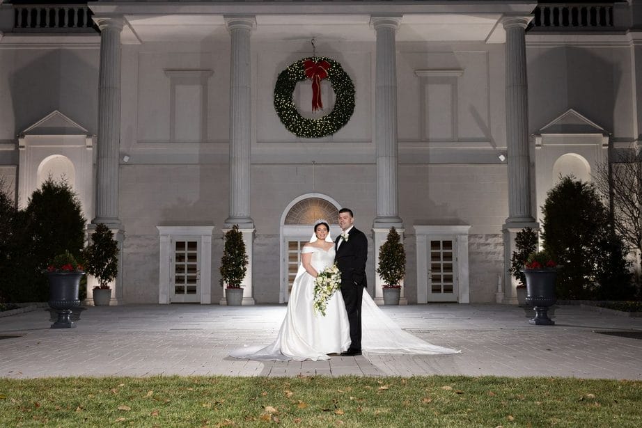 Bride and groom smile happily posing for photos in front of the grand Palace venue with a white stone facade, columns, and a large Christmas wreath