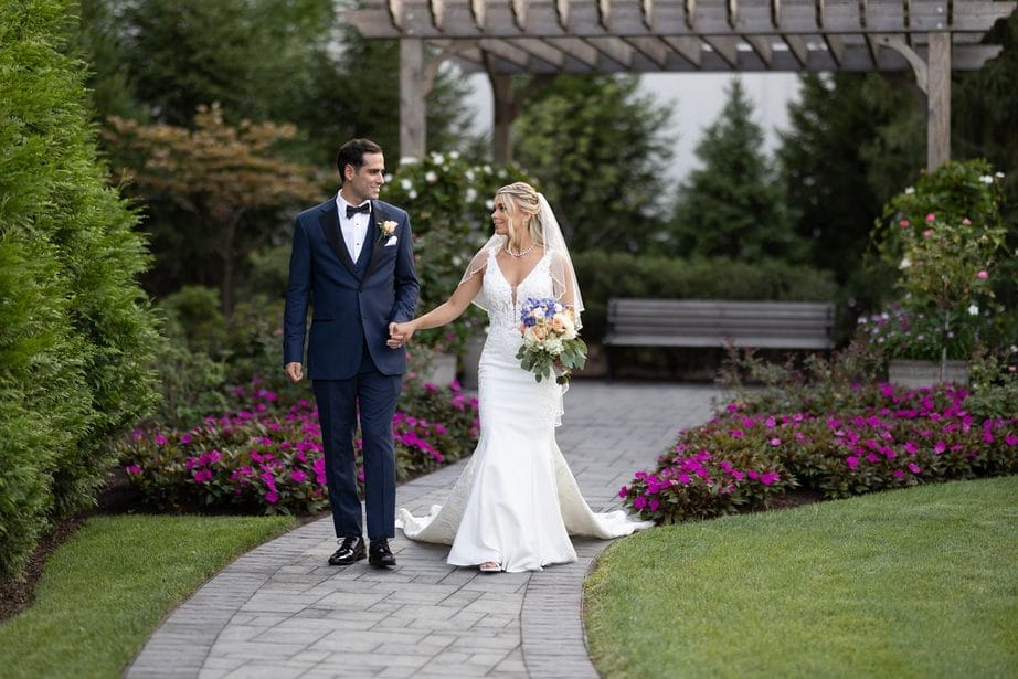 Bride wearing a lace gown and groom wearing a blue suit walk on a brick garden path with purple flowers beside them and a pergola behind them