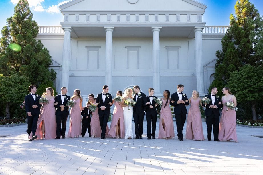 Members of a large wedding party stand in front of the venue's grand white stone facade. The men are wearing black tuxedos and the women are wearing pale pink dresses