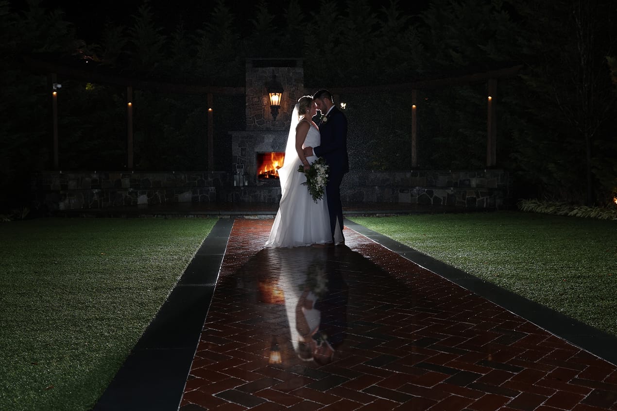 A bride and groom standing in front of a fireplace at night.